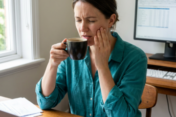 Grace gently presses fingers to her cheek, wincing slightly mid-sip of coffee at her office desk.