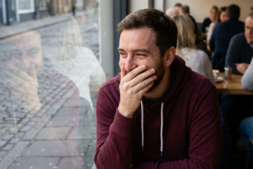 Justin covers his mouth, stifling a laugh at a cafe window seat.